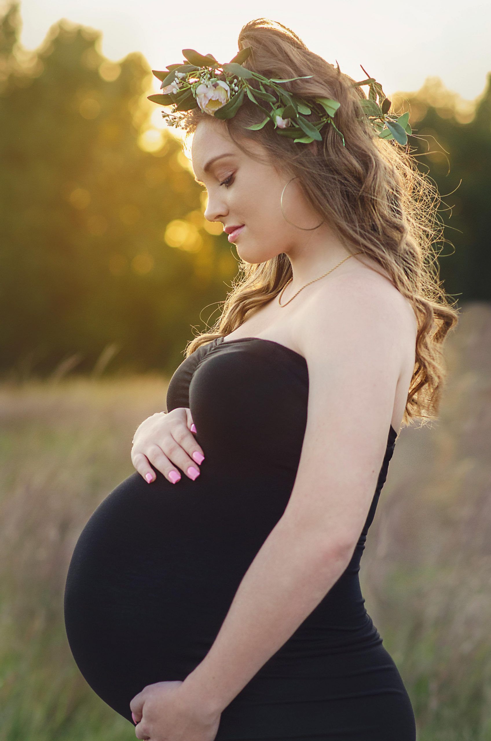 Happy Pregnant woman with hair garland in black dress smiling whilst sun is setting behind trees